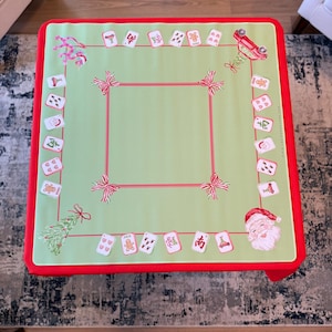 May include: A festive Christmas table cover with a light green central area and a red border. The cover is decorated with illustrations of Santa Claus, gingerbread men, and bows. Designed for a square table, it adds a seasonal touch.