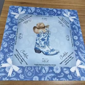 May include: A square fabric mat featuring a blue and white paisley border with white ribbon accents at each corner. The center panel displays a cowboy boot with a blue floral pattern, a tan hat, and instructional text for a line dance called Charleston.