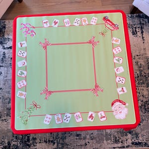 May include: A square, Christmas-themed game table cover with a light green centre and a red border. The cover features a red square border, red bows, and Christmas-themed card illustrations. A Santa Claus illustration is in the corner.