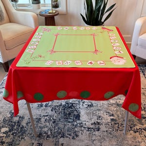May include: A festive Christmas tablecloth. The base is red, with a light green central panel. The green panel has a red border and decorative elements. The red border has green polka dots. Santa Claus and other festive images are printed on the tablecloth.