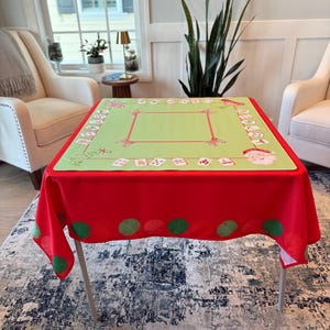 May include: A Christmas-themed tablecloth. The top is light green with red and white decorative elements, including Santa Claus and gift boxes. The tablecloth's border is red with large green polka dots. The table is supported by silver legs.