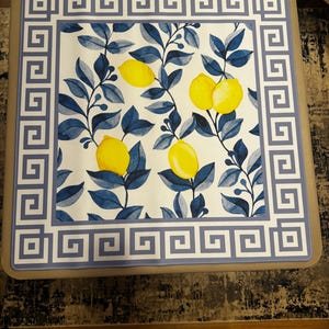 Puede incluir: Mesa de centro cuadrada con una superficie blanca con un dise&ntilde;o de lim&oacute;n y hojas azules. El borde tiene un patr&oacute;n de clave griega azul. La mesa tiene un marco de color neutro.