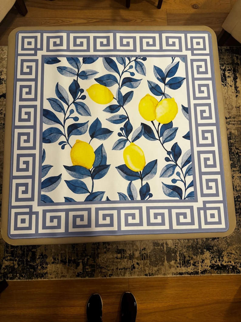 Puede incluir: Mesa de centro cuadrada con una superficie blanca con un dise&ntilde;o de lim&oacute;n y hojas azules. El borde tiene un patr&oacute;n de clave griega azul. La mesa tiene un marco de color neutro.