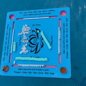 May include: A square blue Pool Jong game board featuring a central black lotus graphic and text. The board includes four slots for tiles, printed rules regarding Jokers and Dragons, and scattered white tiles with black symbols and green tiles.