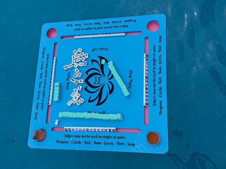 May include: A square blue Pool Jong game board featuring a central black lotus graphic and text. The board includes four slots for tiles, printed rules regarding Jokers and Dragons, and scattered white tiles with black symbols and green tiles.