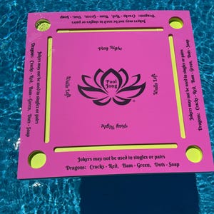 May include: A square pink floating game board with yellow accents and four corner circular cutouts. The center features a black lotus graphic with the text Pool Jong. Text on the edges includes Play Right, Walls Left, and game rules for Jokers and Dragons.