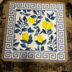 Puede incluir: Mesa de centro cuadrada con una tapa decorativa con un dise&ntilde;o de lim&oacute;n y hojas. El dise&ntilde;o incluye limones amarillos, hojas azules y un fondo blanco. El borde tiene un patr&oacute;n de clave griega azul y blanco.