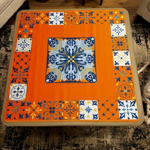 K&ouml;nnte beinhalten: Eine orangefarbene quadratische Tischdecke mit einem zentralen blau-wei&szlig;en Blumendesign. Der Rand weist sich wiederholende geometrische Muster in Blau, Wei&szlig; und Orange auf. Die Tischdecke liegt auf einem beigefarbenen quadratischen Tisch.