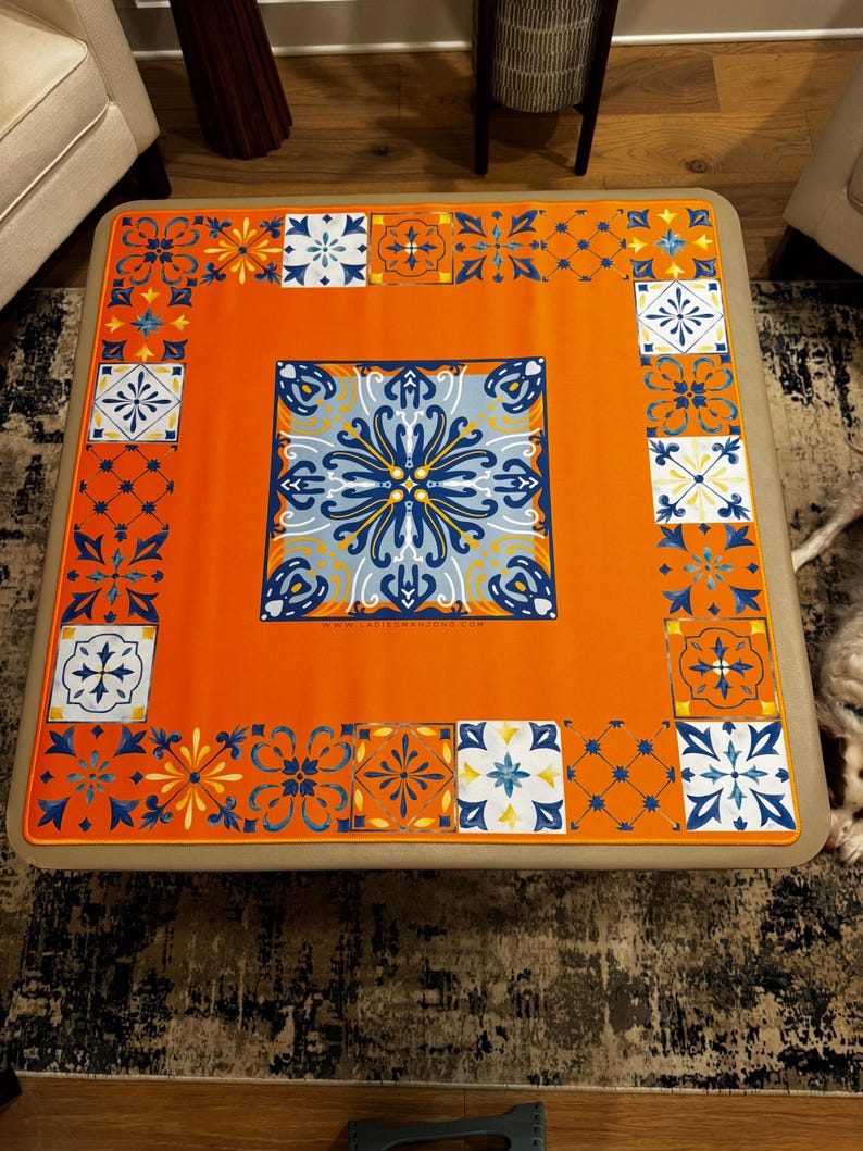 K&ouml;nnte beinhalten: Eine orangefarbene quadratische Tischdecke mit einem zentralen blau-wei&szlig;en Blumendesign. Der Rand weist sich wiederholende geometrische Muster in Blau, Wei&szlig; und Orange auf. Die Tischdecke liegt auf einem beigefarbenen quadratischen Tisch.