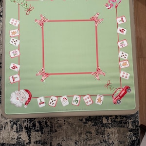 May include: A light green mahjong table cover with a red border and decorative bows. The cover features Christmas-themed card illustrations, including Santa Claus, gingerbread men, and a red car carrying a Christmas tree. The website address is visible.