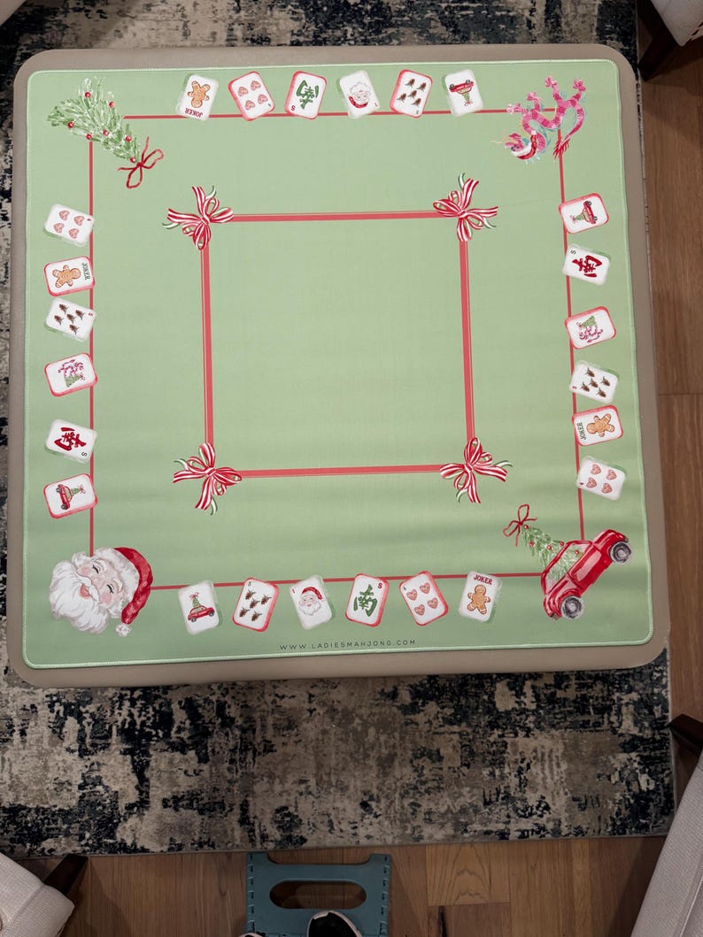 May include: A light green mahjong table cover with a red border and decorative bows. The cover features Christmas-themed card illustrations, including Santa Claus, gingerbread men, and a red car carrying a Christmas tree. The website address is visible.