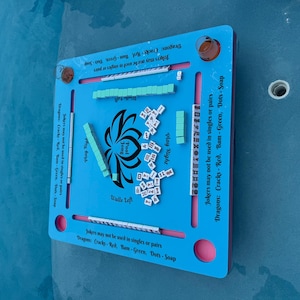 May include: A square, light blue floating game board for Pool Jong with a central lotus graphic. The board features slots for tiles, printed text including "Jokers may not be used in singles or pairs," and "Dragons: Cracks - Red, Bam - Green, Dots - Soap."