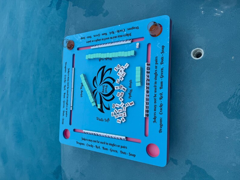 May include: A square, light blue floating game board for Pool Jong with a central lotus graphic. The board features slots for tiles, printed text including "Jokers may not be used in singles or pairs," and "Dragons: Cracks - Red, Bam - Green, Dots - Soap."