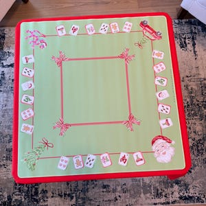 May include: A festive Christmas table cover with a light green central area and a red border. The cover is decorated with illustrations of Santa Claus, gingerbread men, and Christmas tree designs. Red ribbons and card illustrations are also present.