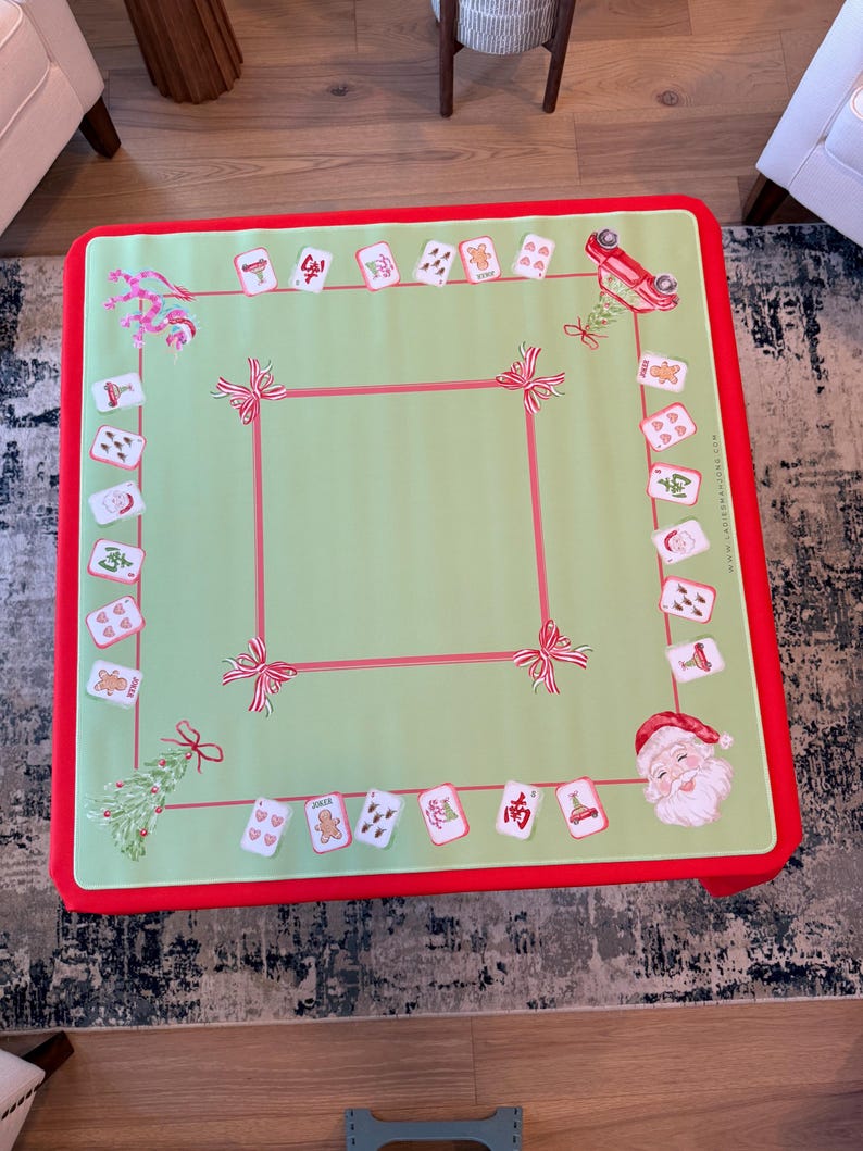 May include: A festive Christmas table cover with a light green central area and a red border. The cover is decorated with illustrations of Santa Claus, gingerbread men, and Christmas tree designs. Red ribbons and card illustrations are also present.