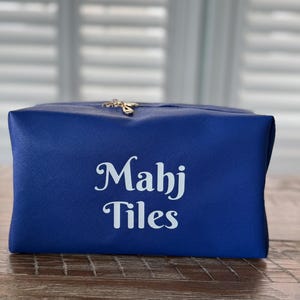 May include: A royal blue cosmetic bag with the text "Mahj Tiles" in white script. The bag has a gold-colored zipper pull and a textured surface. The bag is rectangular and sits on a wooden surface.