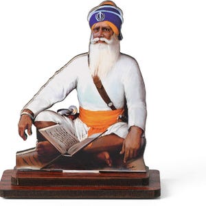 May include: A wooden figurine of a Sikh man in traditional attire, seated and reading a book. He wears a white tunic, orange sash, and a blue and white turban. The figurine is mounted on a dark wooden base.