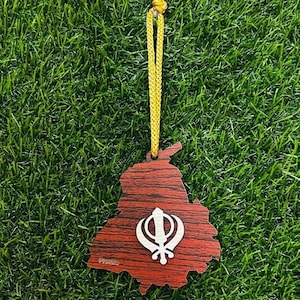 May include: A wooden ornament in the shape of a map of Punjab, with a white Khanda symbol. The ornament is red with a wood grain pattern and hangs from a yellow cord. The background is green.