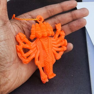 May include: Bright orange figurine of a deity with multiple arms and heads, held in a hand. The figurine is made of plastic and has a loop at the top with a yellow string attached. The figure is detailed with intricate carvings.