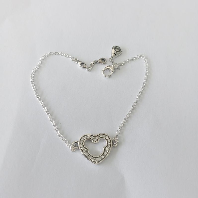 Peut inclure: Un bracelet en argent avec un charm en forme de c&oelig;ur int&eacute;grant une silhouette de Mickey Mouse. Le charm est serti de petits pierres pr&eacute;cieuses transparentes. Le bracelet a une cha&icirc;ne fine et une fermeture &agrave; mousqueton.