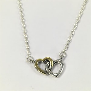 Può includere: Una collana d'argento con due ciondoli a forma di cuore intrecciati. Un cuore è dorato, l'altro è argentato. La catena è un delicato design a maglie d'argento, adatto all'uso quotidiano.