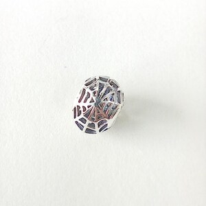 May include: A silver bead featuring a spiderweb design. The oval-shaped bead has a detailed web pattern across its surface, creating a delicate, open structure. This intricate design suggests it's intended for jewellery making, possibly for bracelets or necklaces.