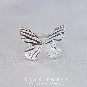 May include: A silver butterfly ring with detailed wing patterns. The ring's design features open wings, creating a delicate and elegant look. The ring is set against a soft, neutral background, highlighting the jewellery's craftsmanship.