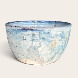 May include: A ceramic bowl featuring a reactive glaze with blended shades of light blue, white, and hints of tan. The exterior surface displays a smooth, glossy finish with subtle vertical streaks and a darker blue rim around the top edge.