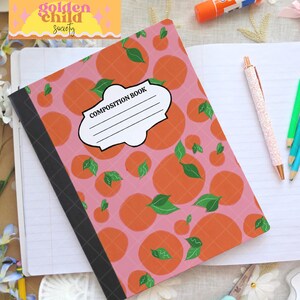 May include: A composition book with a pink cover decorated with orange fruit and green leaves. The cover has a white label with the words "COMPOSITION BOOK". The book is open to a blank page, with a pen and pencils nearby.