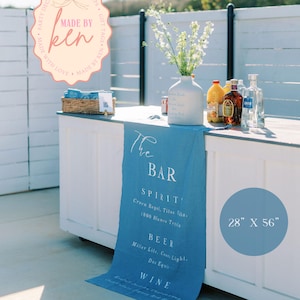 May include: A blue bar runner with white text that reads "The Bar" and lists drink options. The runner is 28 inches by 56 inches. A white bar with bottles, a vase of flowers, and a sign that says "Made by Ken" are also visible.