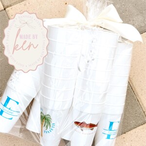 May include: A bundle of white plastic cups wrapped in clear cellophane and tied with a white bow. Some cups feature a blue initial and name, a palm tree with the word "PARADISE", and a fish illustration with text.