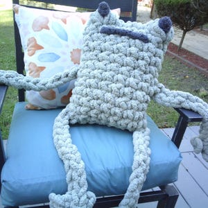 May include: A large, handmade, light green frog-shaped pillow with long arms and legs. The frog has dark gray accents for the eyes and mouth. It is sitting on a blue cushion on a black chair, with a decorative pillow behind it.