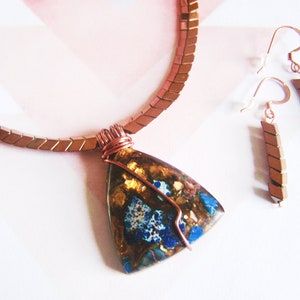 May include: A copper necklace with a triangular pendant featuring a blue and gold stone. The pendant is wrapped in copper wire. The necklace is paired with matching copper earrings.