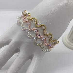 Può includere: Tre delicati braccialetti in argento, oro rosa e oro. Ogni braccialetto presenta un design ondulato con perline chiare e iridescenti. I braccialetti sono esposti su una forma a mano bianca, che ne mette in risalto i dettagli.