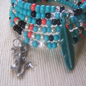 Op de afbeelding: Meerstrengige turquoise, zwarte, zilveren en koraal kralen armband met een turquoise veren bedel en een zilveren hagedis bedel.