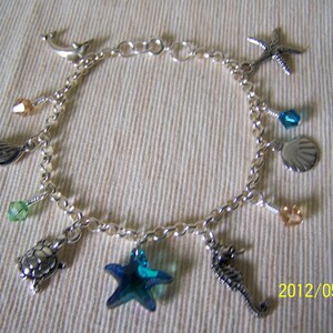 Könnte beinhalten: Ein silbernes Charm-Armband mit verschiedenen Meeres-Anhängern, darunter eine Muschel, ein Seestern, ein Seepferdchen und eine Schildkröte. Das Armband enthält auch blaue und grüne Kristallperlen.
