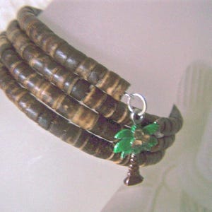 May include: A wrap bracelet made of dark brown, cylindrical beads. A small, green and brown palm tree charm hangs from a silver-colored ring. The bracelet is wrapped around a wrist.