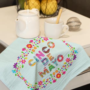 May include: A pale blue Cinco de Mayo tea towel with a colourful floral border. The towel displays the words "Cinco de Mayo" in a vibrant, patterned font. A white teapot, cup, and fruit basket are in the background.