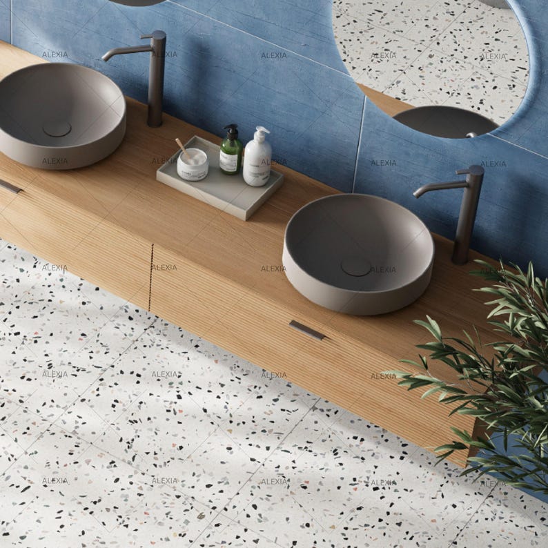 May include: A modern bathroom vanity with two gray bowl sinks, chrome faucets, and a wooden countertop. The vanity includes a small tray with toiletries. The background features blue tile and a round mirror. The floor is white with speckled patterns.