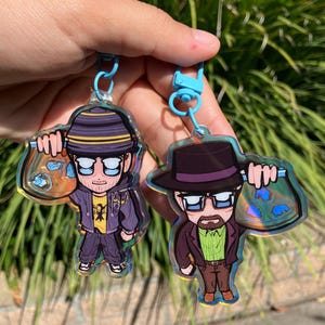 以下が含まれることがあります： 2つのアクリルキーホルダー。紫と黄色の服を着たキャラクターがバッグを持っています。もう一方のキーホルダーは、茶色のスーツと帽子をかぶったキャラクターがバッグを持っています。どちらのキーホルダーも青色の金具が付いています。