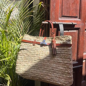 May include: A large, rectangular woven straw tote bag with brown leather detailing. The bag has a braided leather handle, a leather strap with a silver buckle, and a decorative tassel. The bag is hanging on a metal bar in front of a wooden door.
