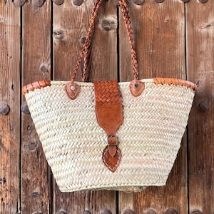 Peut inclure: Un sac fourre-tout en paille tressée avec une poignée et des accents en cuir marron. Le sac présente un motif tissé, un rabat en cuir avec une fermeture à boucle et une poignée tressée. Le sac est posé sur un fond en bois.