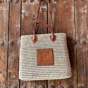 Peut inclure: Sac fourre-tout en paille tressée avec anses en cuir marron et un patch en cuir avec la lettre "E". Le sac est de forme rectangulaire et est posé sur un fond de porte en bois.
