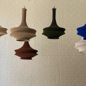 Puede incluir: Un conjunto de lámparas colgantes en varios colores, incluyendo beige, marrón, verde oliva, marrón oscuro, azul y blanco. Las lámparas tienen una textura tejida y un diseño escalonado único, creando una luminaria decorativa.