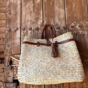 May include: Woven straw tote bag with brown leather detailing. The bag has a braided handle, a leather strap, and a decorative tassel. It is set against a rustic wooden door.