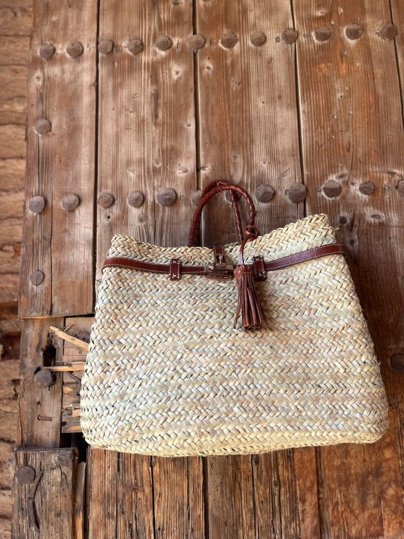 May include: Woven straw tote bag with brown leather detailing. The bag has a braided handle, a leather strap, and a decorative tassel. It is set against a rustic wooden door.