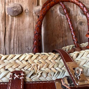 May include: A close-up of a woven straw tote bag with brown leather detailing. The bag has a braided leather handle, leather straps, and a metal clasp. The bag is set against a rustic wooden background.