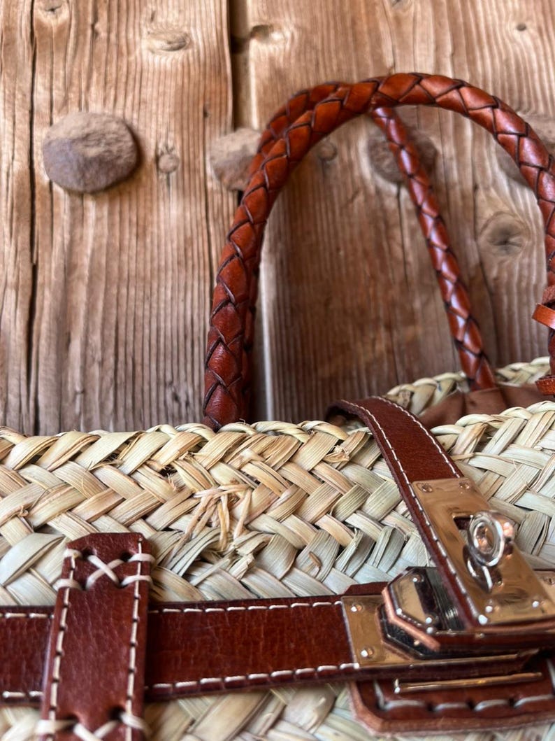 May include: A close-up of a woven straw tote bag with brown leather detailing. The bag has a braided leather handle, leather straps, and a metal clasp. The bag is set against a rustic wooden background.