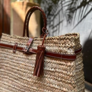 May include: Woven straw tote bag with brown leather detailing. It has a braided handle, a leather strap with a buckle, and a decorative tassel. The bag is designed with a spacious interior.