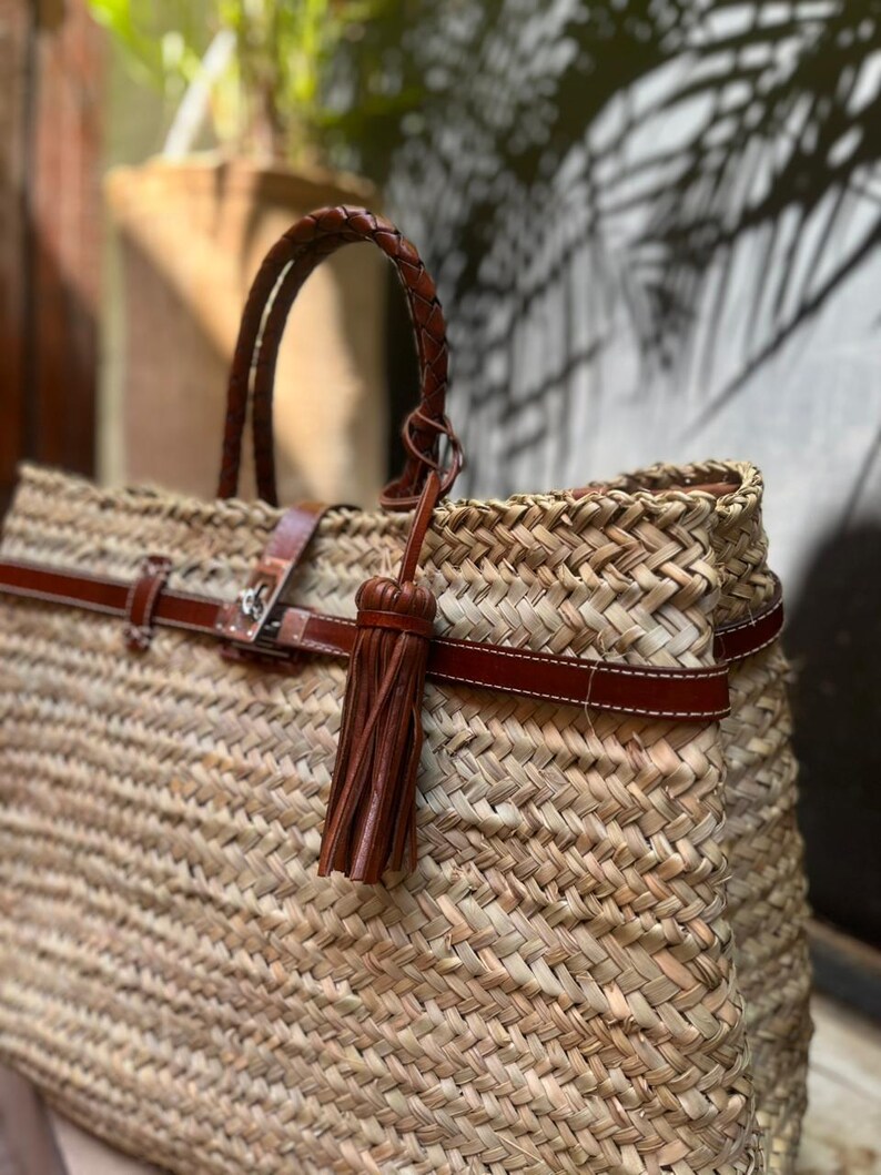 May include: Woven straw tote bag with brown leather detailing. It has a braided handle, a leather strap with a buckle, and a decorative tassel. The bag is designed with a spacious interior.